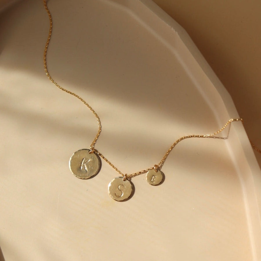 14k gold fill Monogram coin necklace on a peach colored plate in the sunlight.