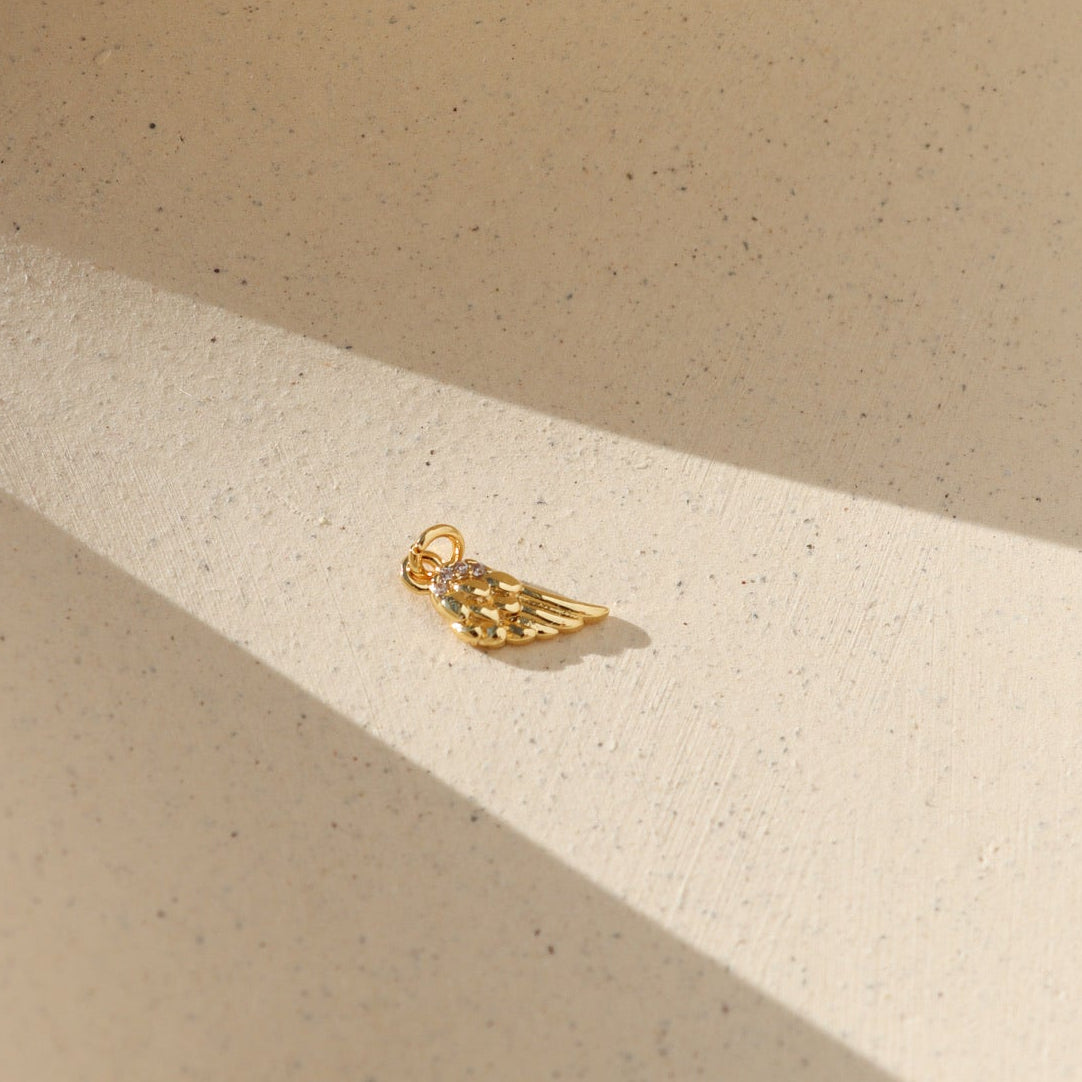 Gold angel wing charm with sparkling crystal details, photographed on a warm neutral surface with soft natural light and shadows.