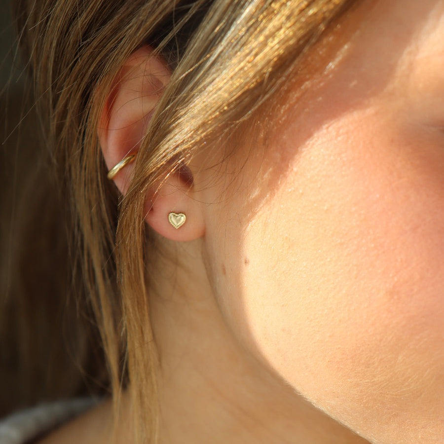 dainty gold heart earring with a patterned border, pictured on a model in sunlight