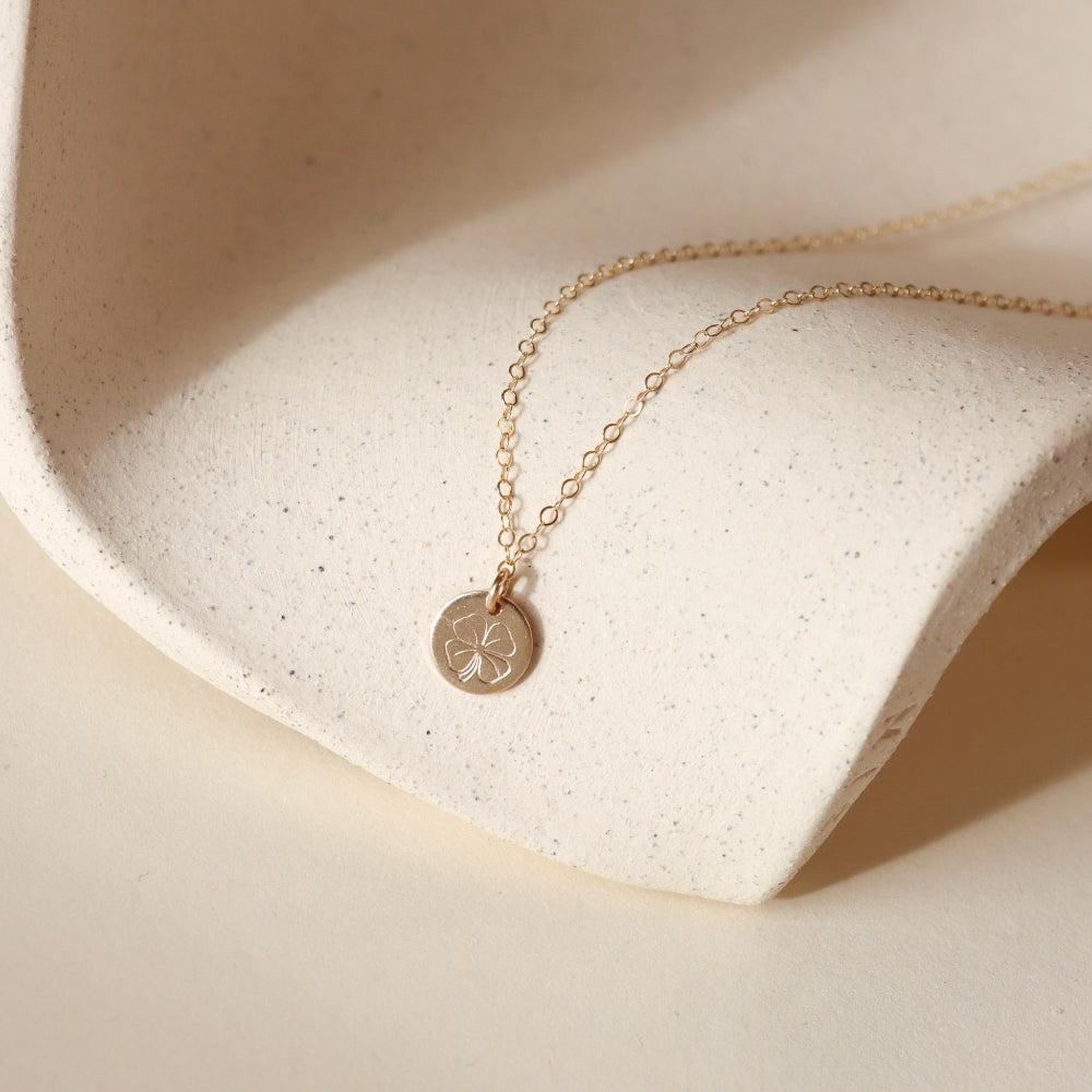 Gold filled necklace with a round hand stamped four leaf clover pendant on a beige stone surface