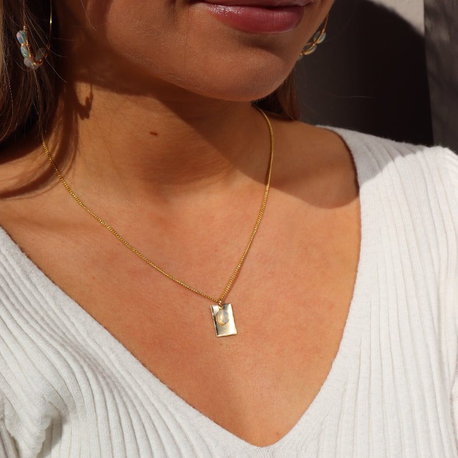 Model wearing 14k gold fill Everly Charm necklace. This necklace feature a simple chain with a box charm and the charm has hammering on the outside, Laid on top of the box charm is a opal stone.