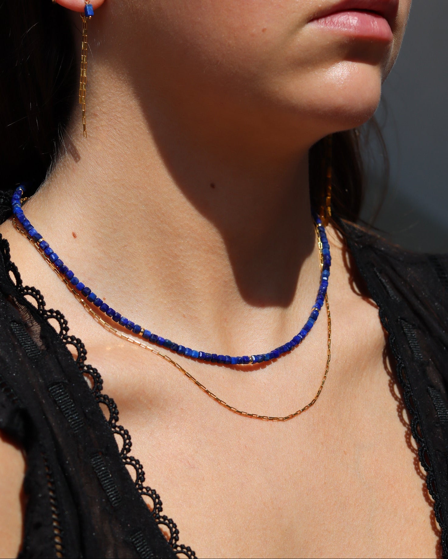 A woman wearing a deep blue beaded necklace layered with a thin gold chain, matching gold drop earrings with a blue accent, and a black lace top.