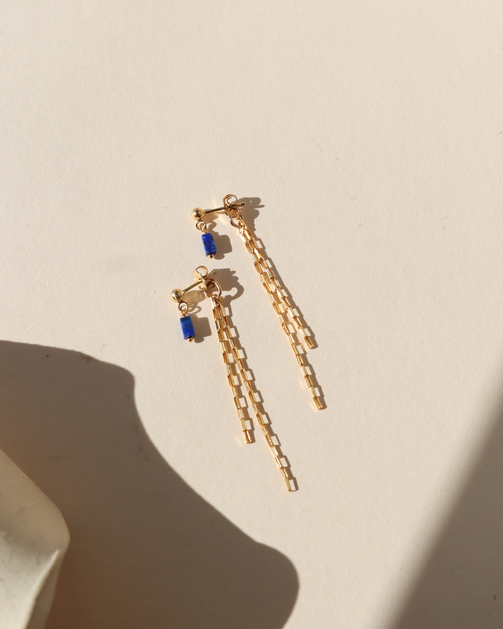 Gold stud earrings with a small rectangular blue stone dangle and long, layered chain fringe lying on a beige surface