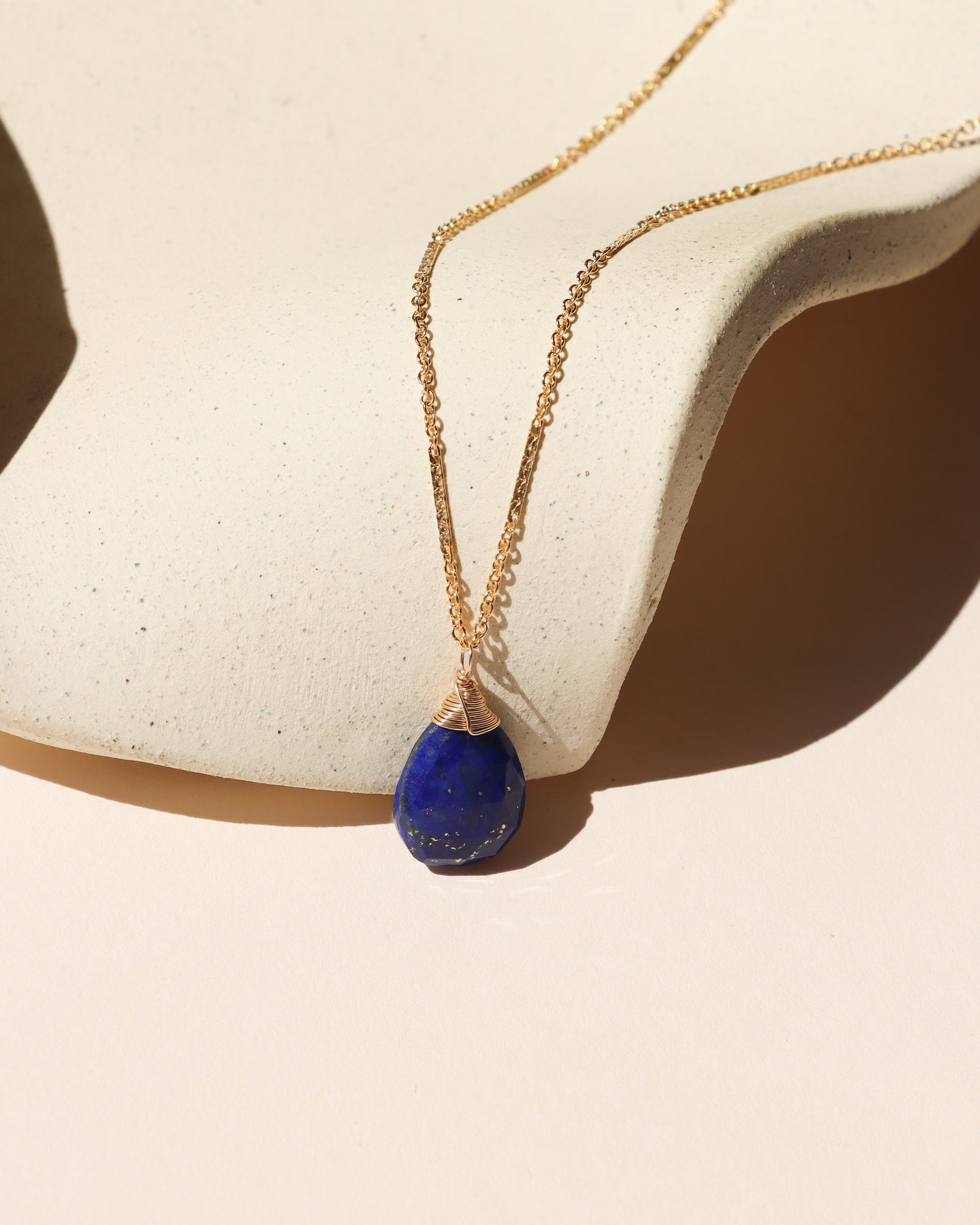 Gold chain necklace with a deep blue lapis lazuli pendant displayed on a cream stone surface in warm sunlight.