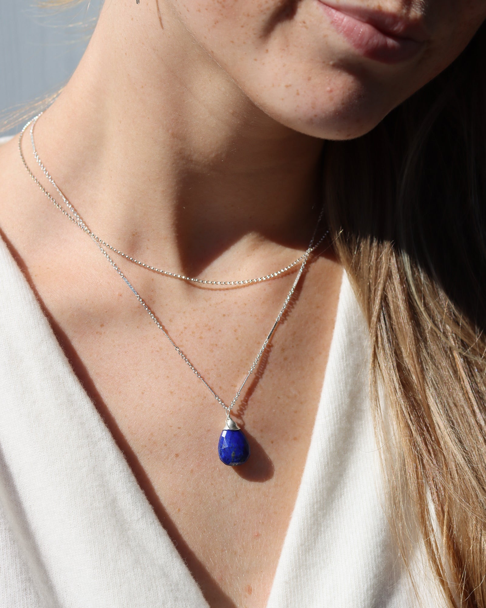 Close-up of a woman wearing a layered silver necklace with a teardrop-shaped lapis lazuli pendant against a white top.