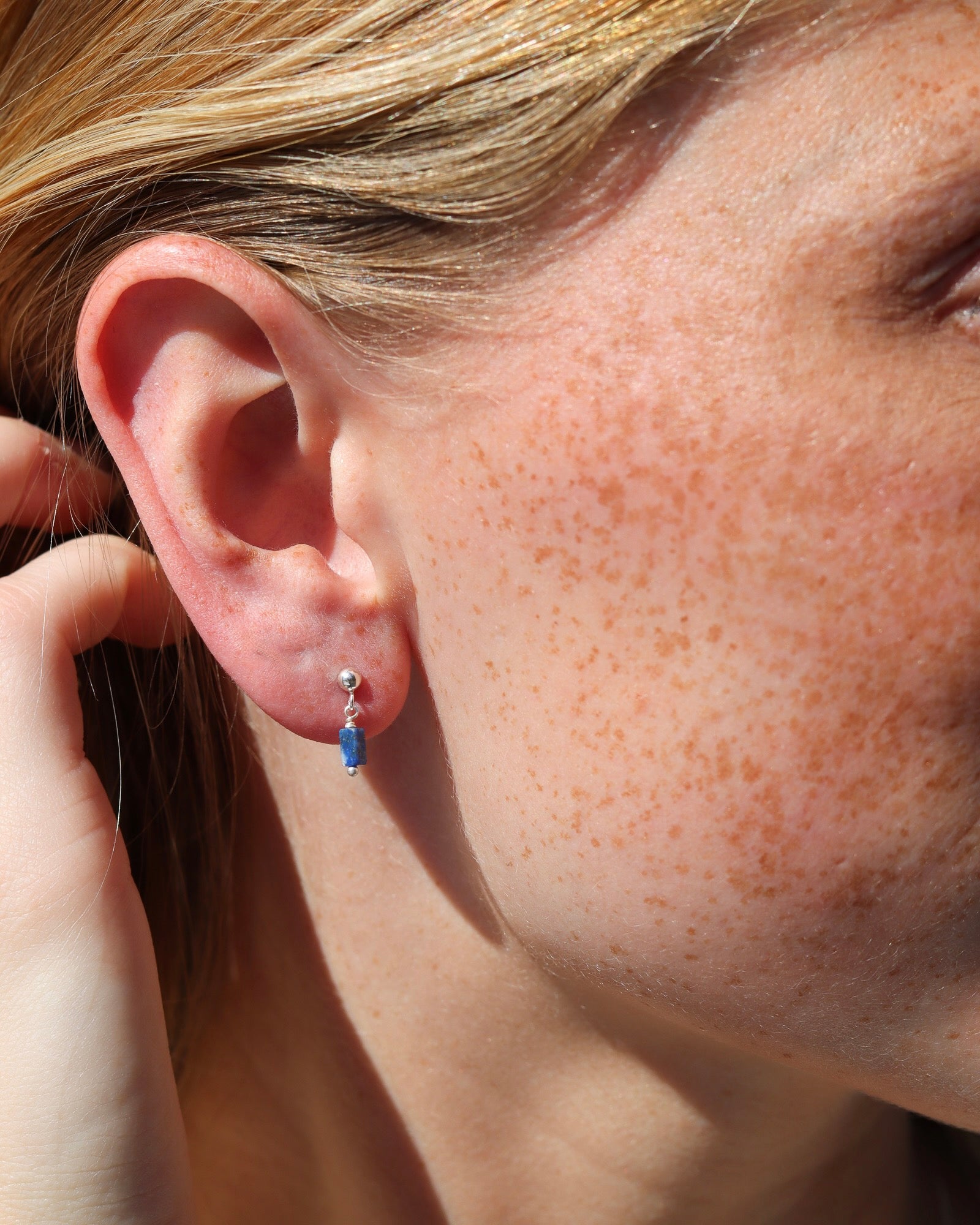 Close-up of a person wearing a silver stud earring with a small, rectangular blue stone dangle.