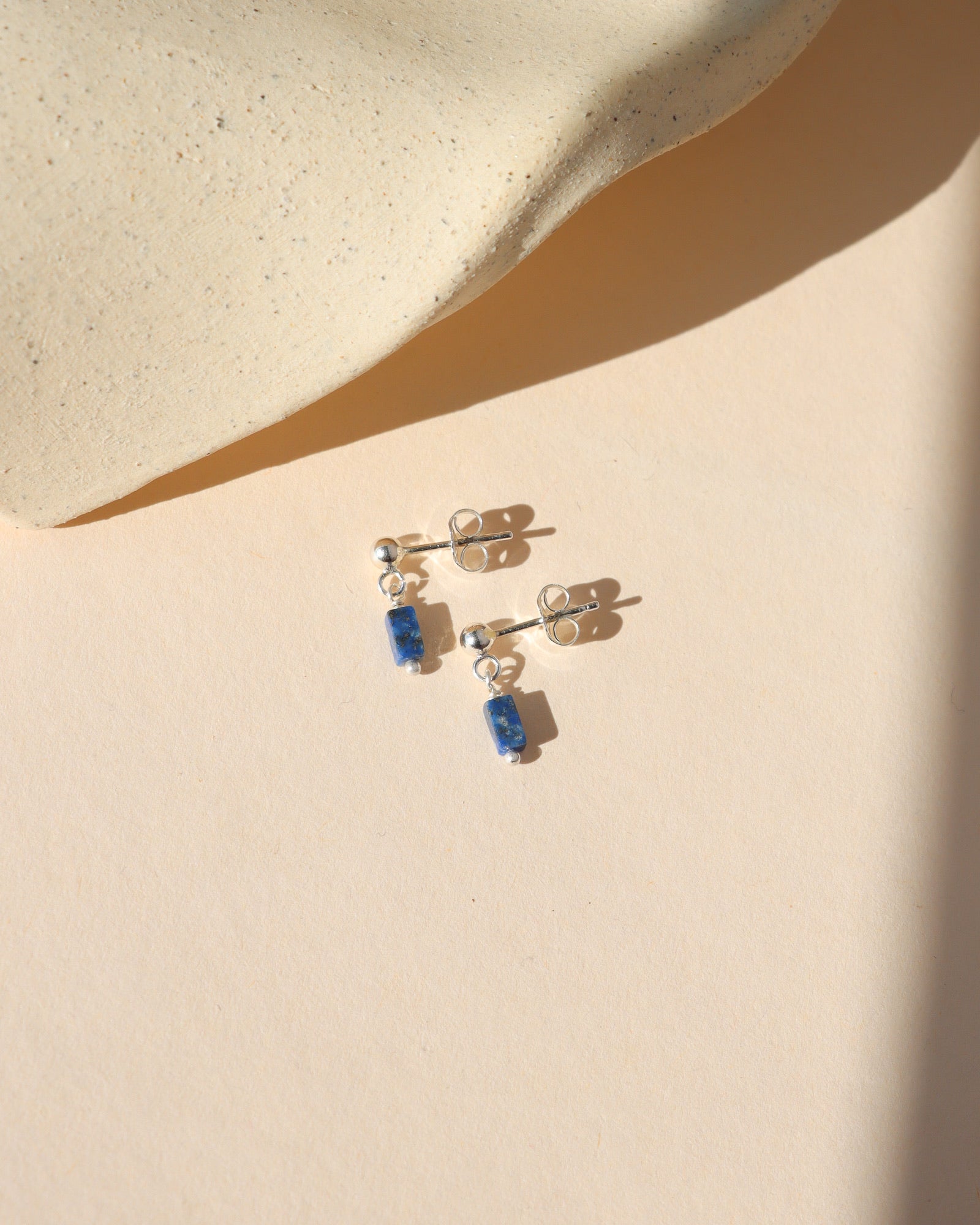 A pair of silver stud earrings featuring small, rectangular blue stone dangles set against a neutral beige background.