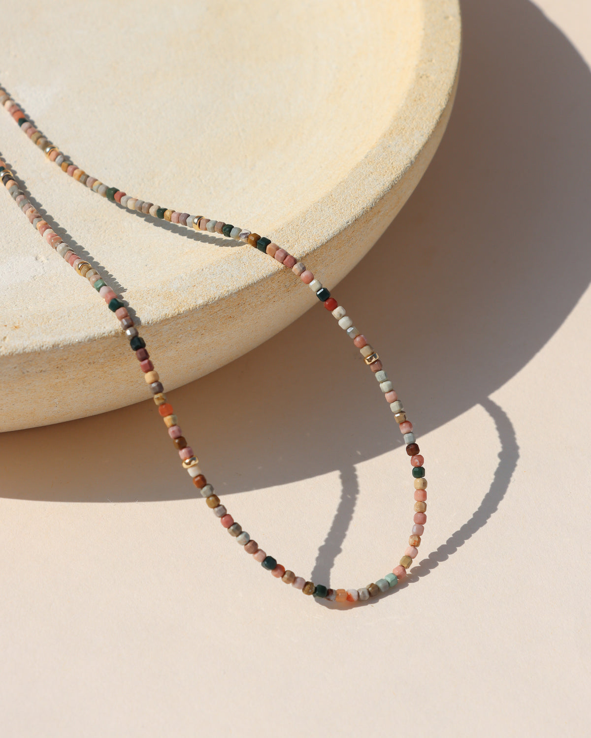 Multicolored gemstone beaded necklace with tiny square beads in blush pink, sage green, cream, rust, and earthy tones, accented with small gold beads and styled on a cream display.
