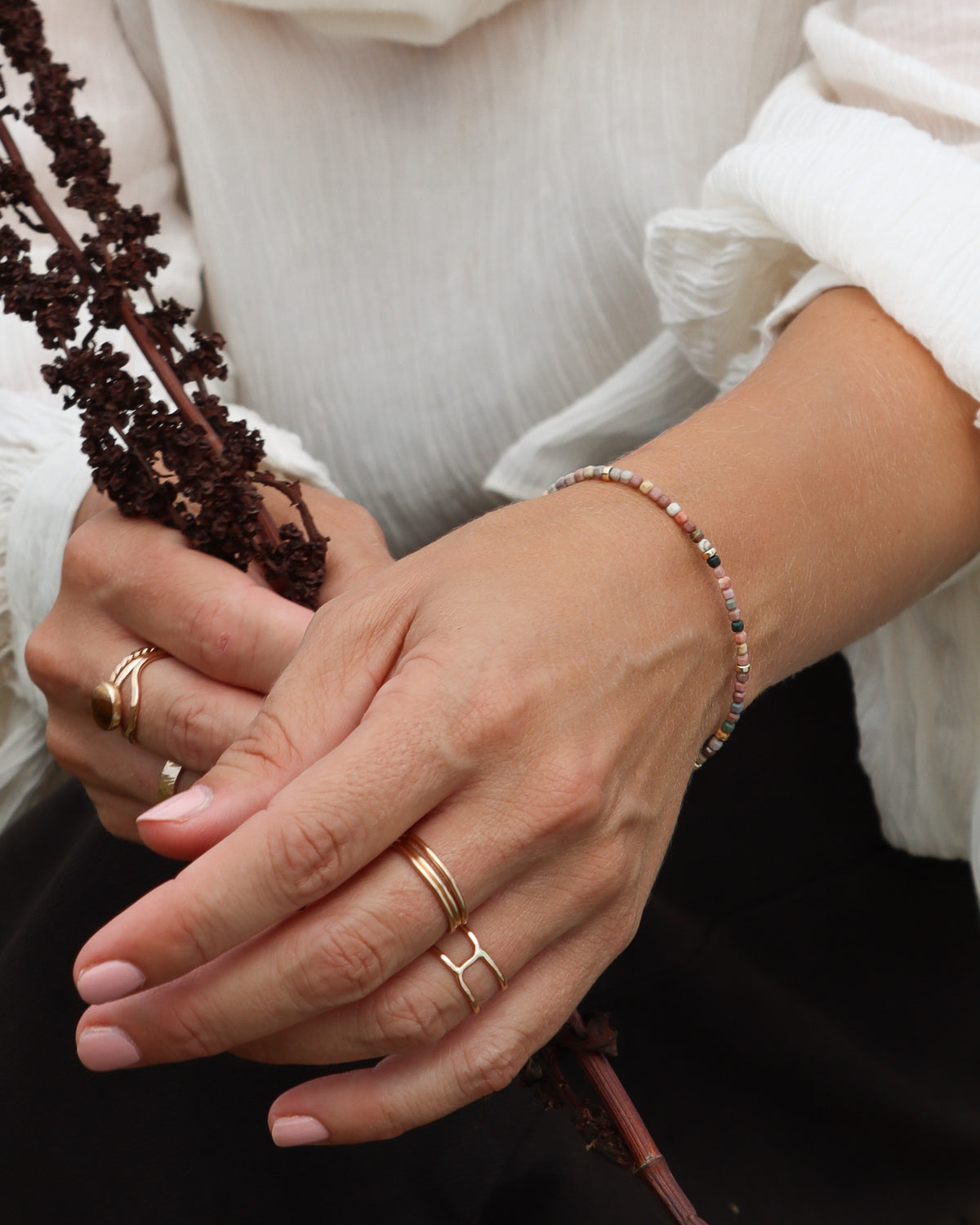Hands wearing layered gold rings and a delicate multicolored beaded bracelet, styled with a white blouse.