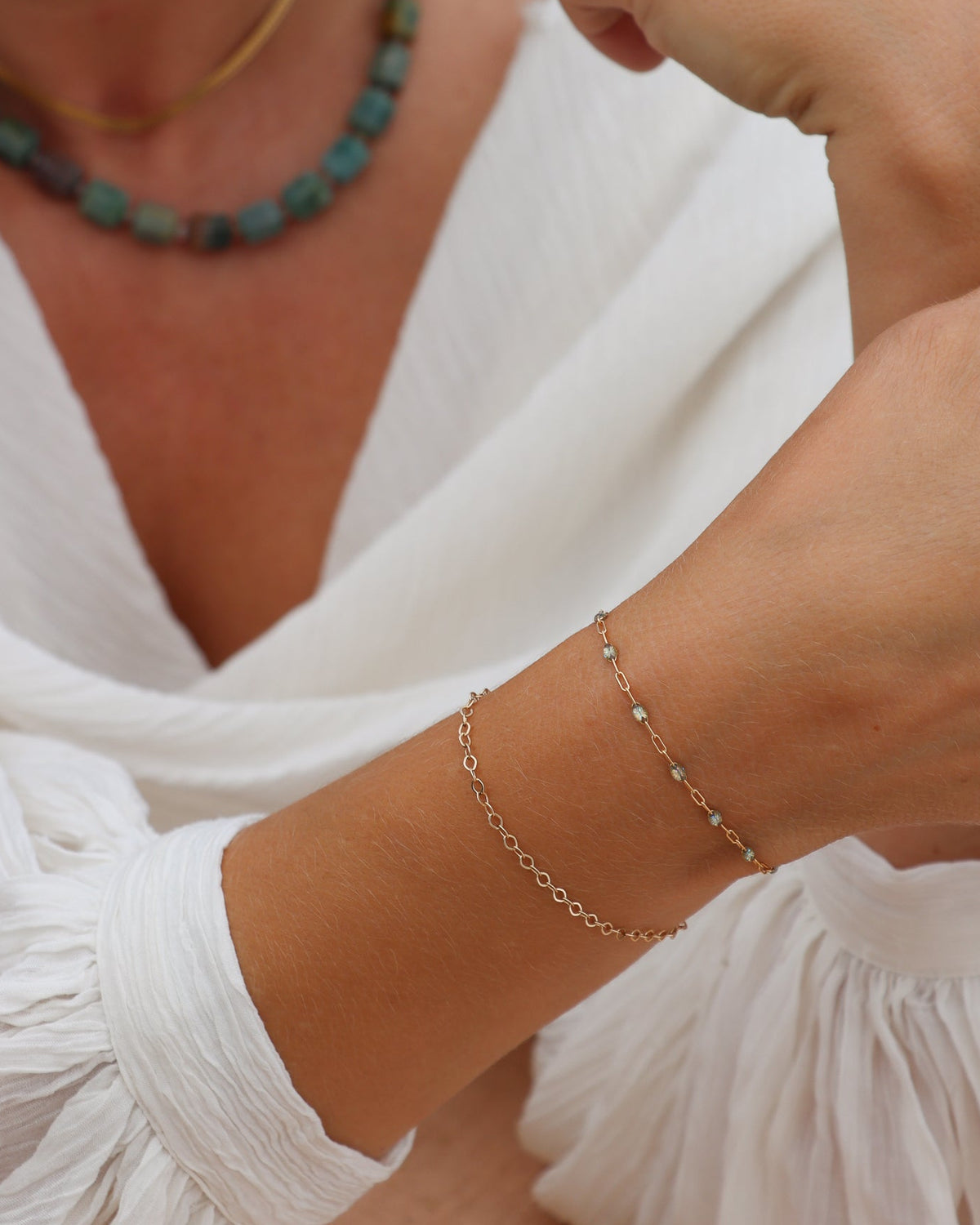 Close-up of layered gold chain bracelets, including a delicate bracelet with small blue-green accents, styled with a white blouse.