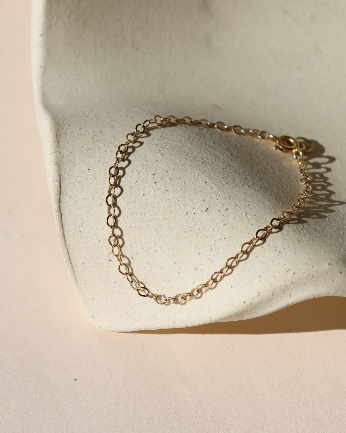 Delicate gold oval-link chain bracelet displayed on a cream ceramic surface in warm natural sunlight.