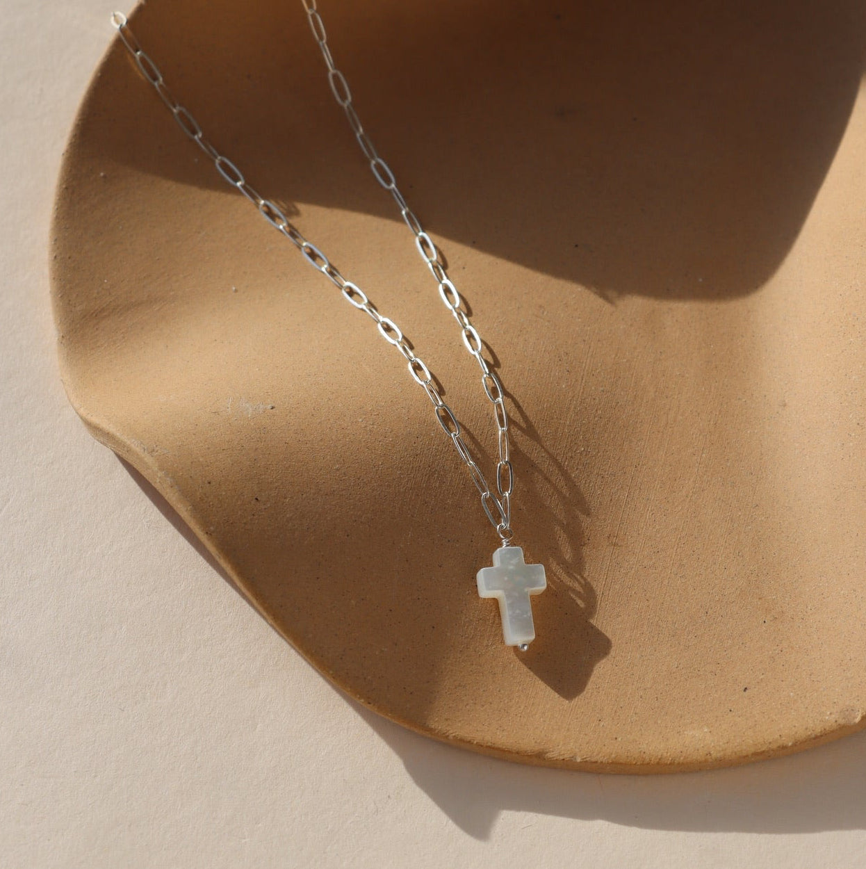 sterling silver paperclip chain necklace with mother of pearl cross pendant charm on a tan dish