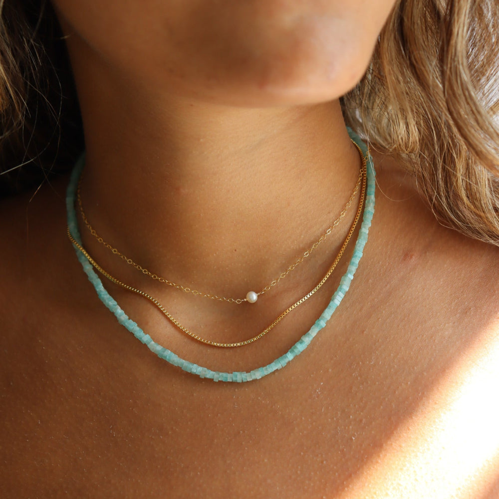 Model wearing a styled necklace stack. Featuring a simple 14k gold fill chain with a mini pearl anchored in the center. It is paired with a rolo 14k gold fill chain, and a fully beaded gemstone necklace to create a three necklace stack. 