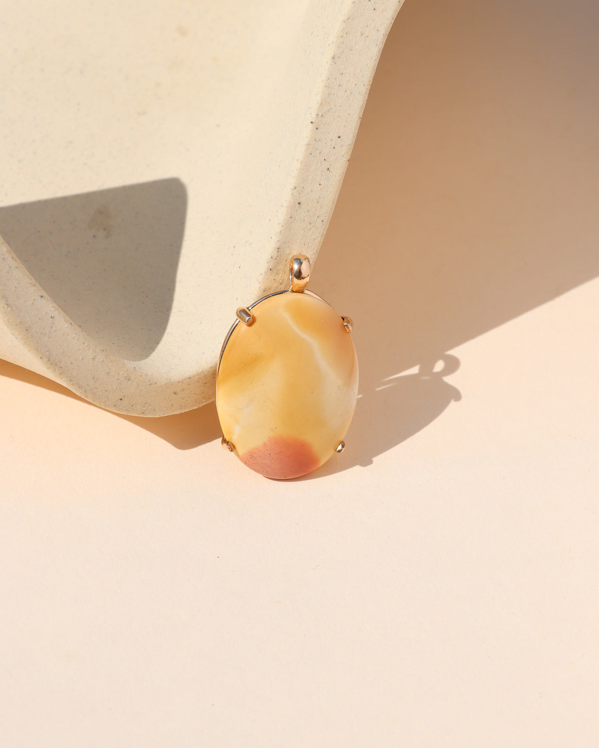 Oval golden yellow and cream stone pendant with a soft red accent, set in gold and displayed beside a textured cream ceramic form.