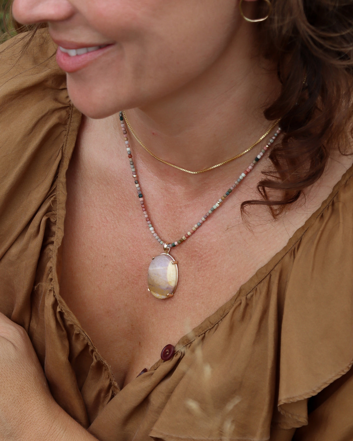Oval cream, lavender, and golden stone pendant on a multicolored beaded necklace, layered with a delicate gold chain and worn with a brown top.