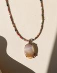 Multicolored beaded necklace featuring an oval cream, lavender, and golden stone pendant in a gold setting, photographed in warm sunlight.
