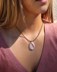 Oval cream and red stone pendant on an olive cord necklace, worn with a plum top in warm sunlight.