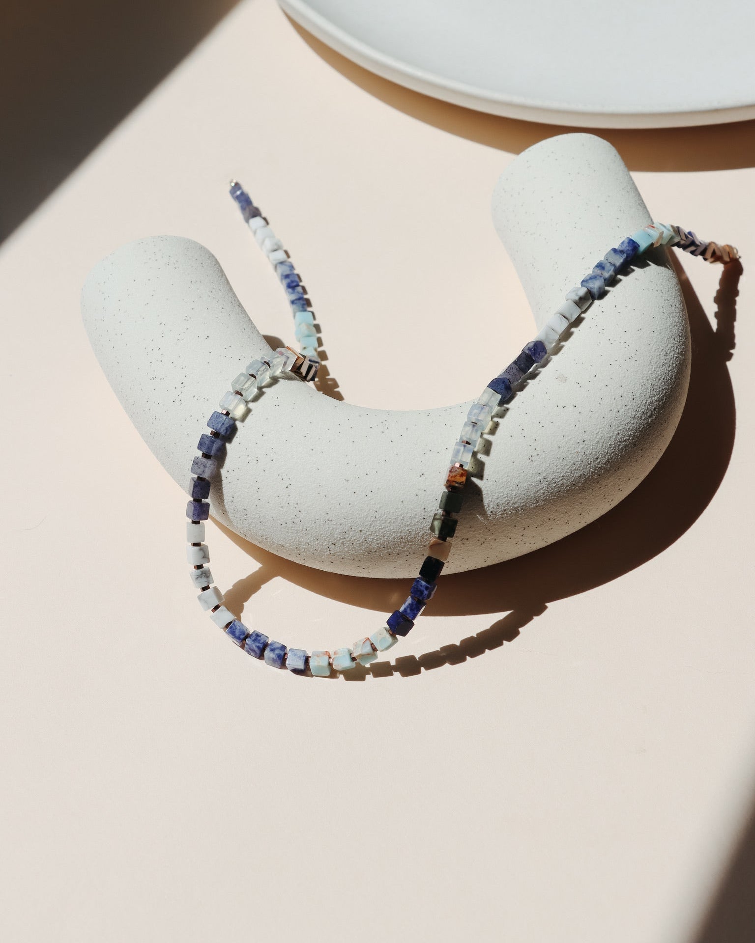 Beaded necklace draped on a beige arc dish in the sunlight.