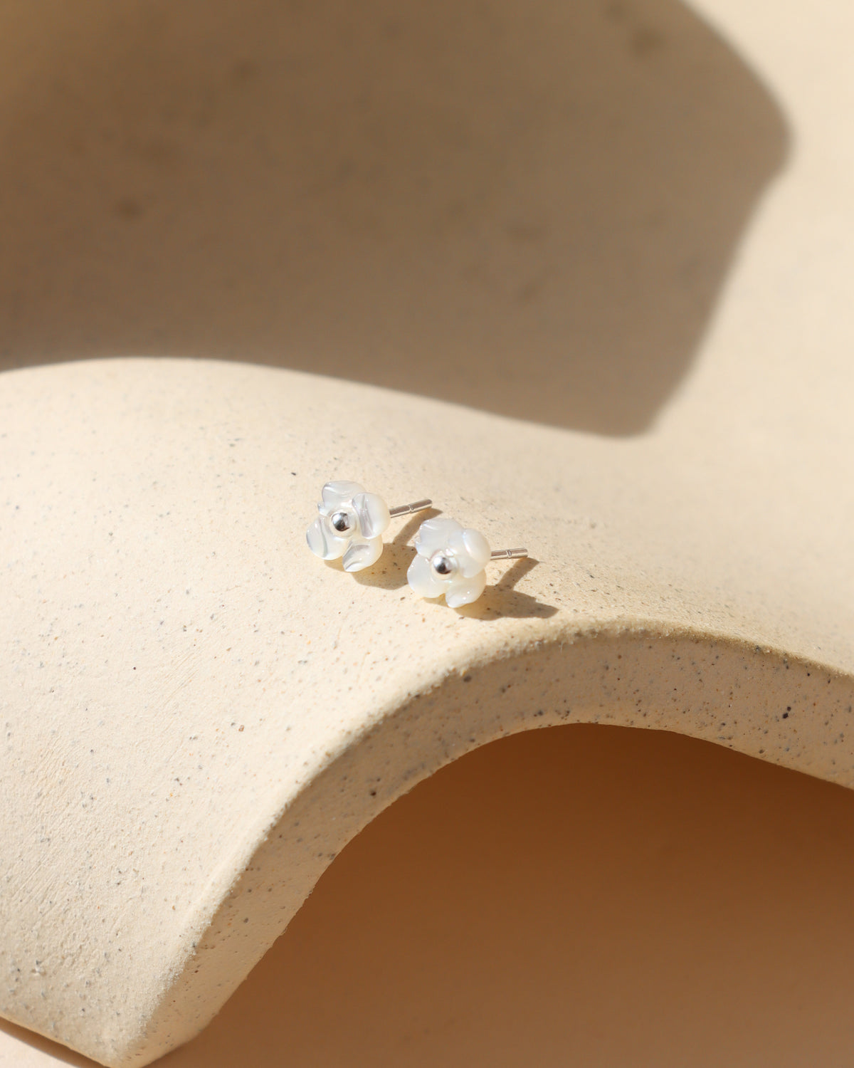 Pair of sterling silver floral stud earrings featuring delicate white mother-of-pearl petals and silver centers, resting on a curved cream stone surface in warm natural sunlight.