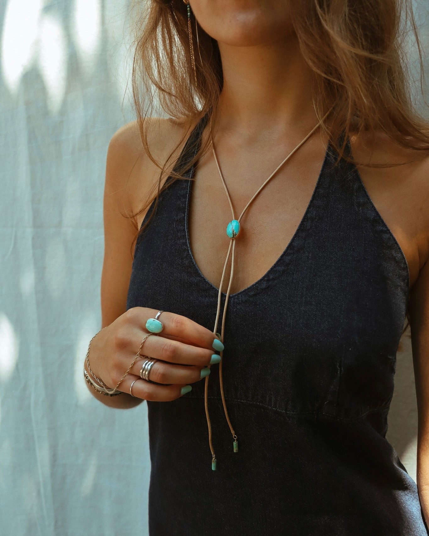 Model wearing a turquoise bolo necklace with natural leather cord and a genuine turquoise slide, styled with a turquoise statement ring, layered silver rings, and delicate bracelets.