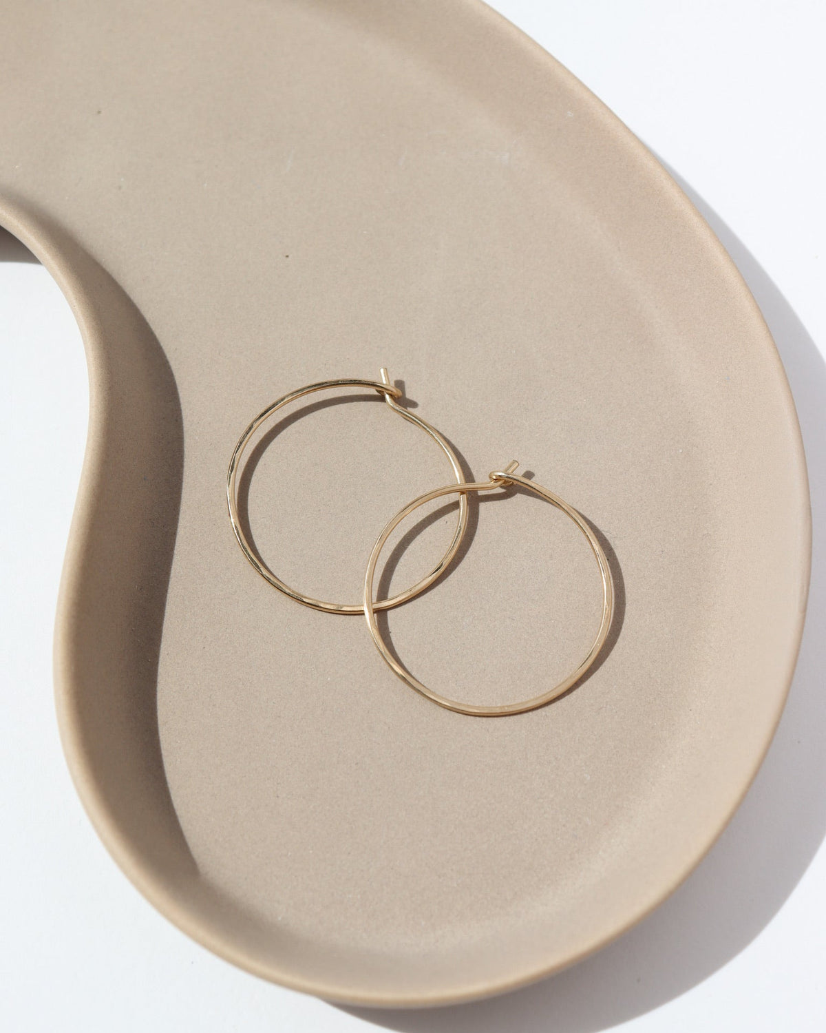 14k solid gold Organic Hoops by Token Jewelry. The hoops are a circle shape with hammered solid gold wire, photographed on a tan ceramic dish