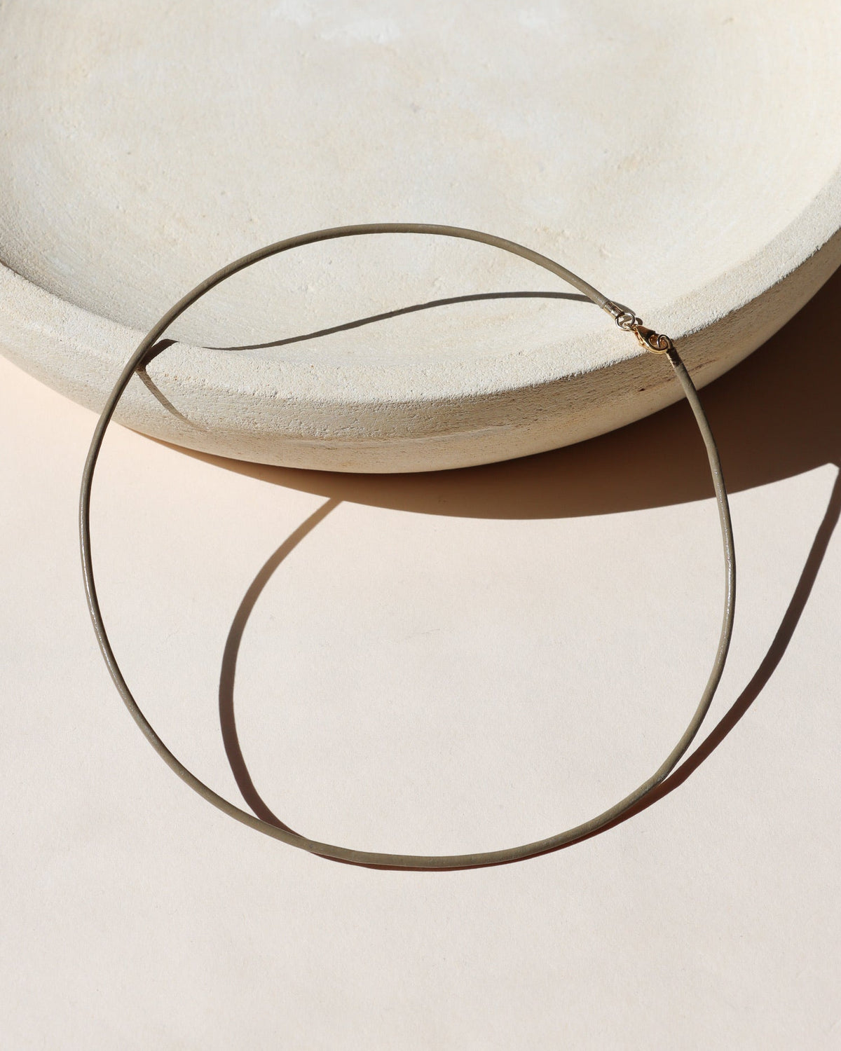 Thin sand leather cord necklace with a small gold clasp, arranged in a circle beside a textured cream ceramic form in warm sunlight.
