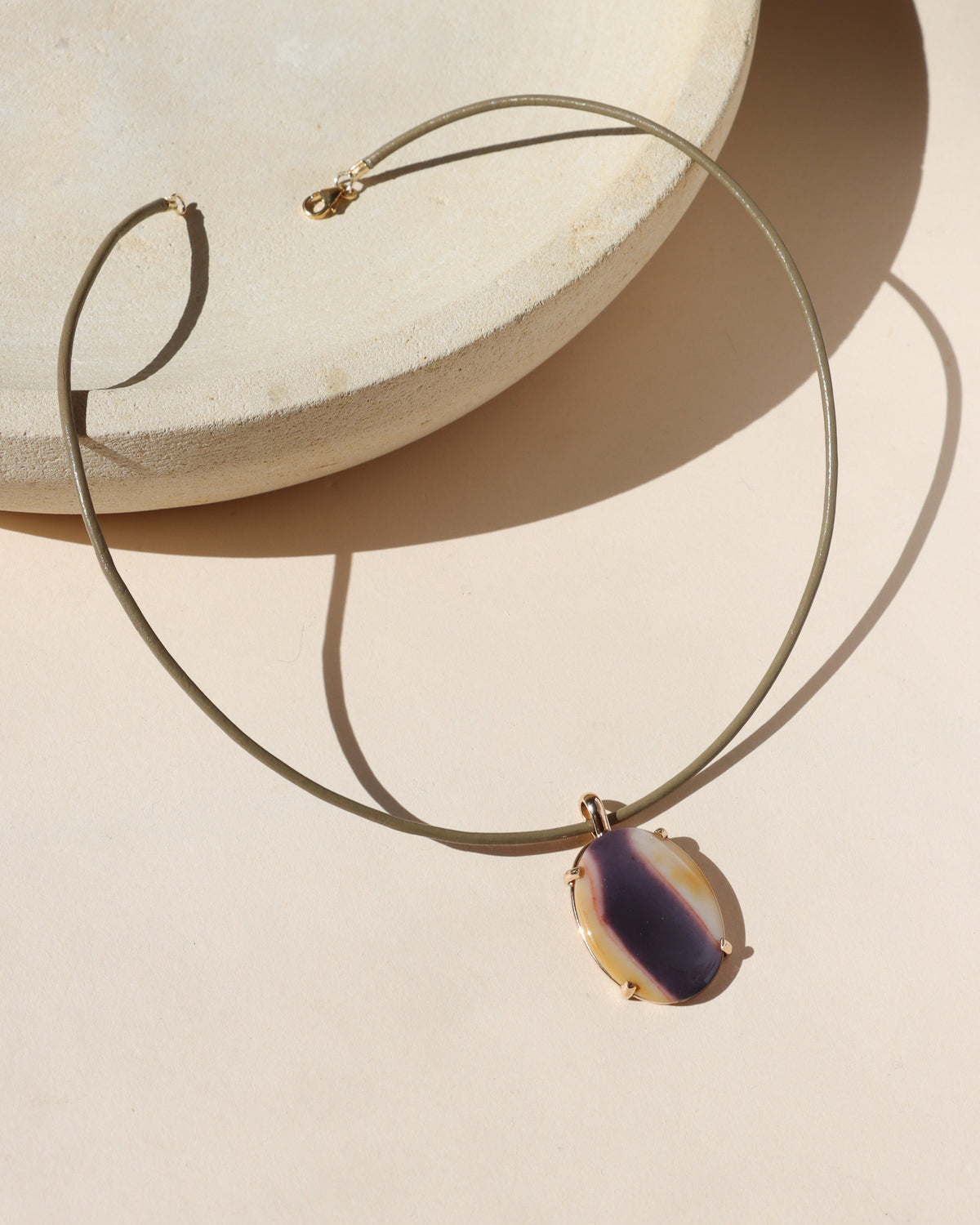 Sand leather cord necklace featuring an oval purple, cream, and golden stone pendant in a gold setting, photographed in warm sunlight.