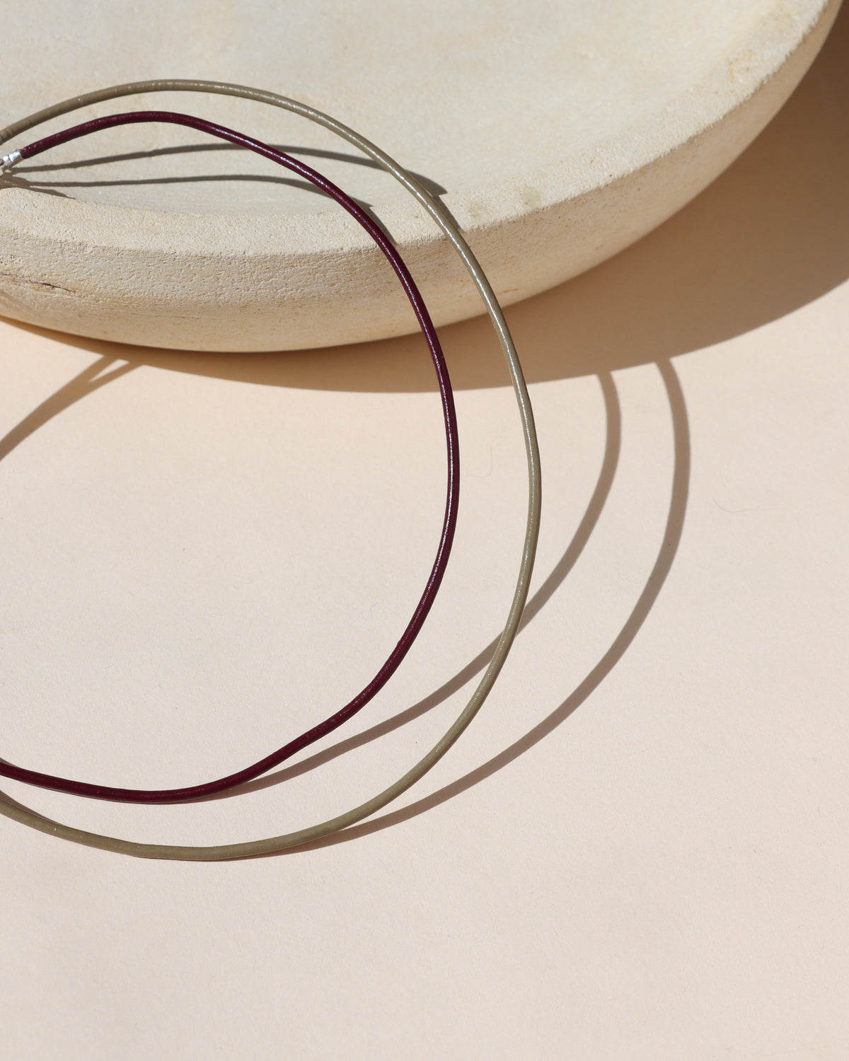 Sand and plum leather cord necklaces draped over a textured cream ceramic form, casting curved shadows in warm sunlight.