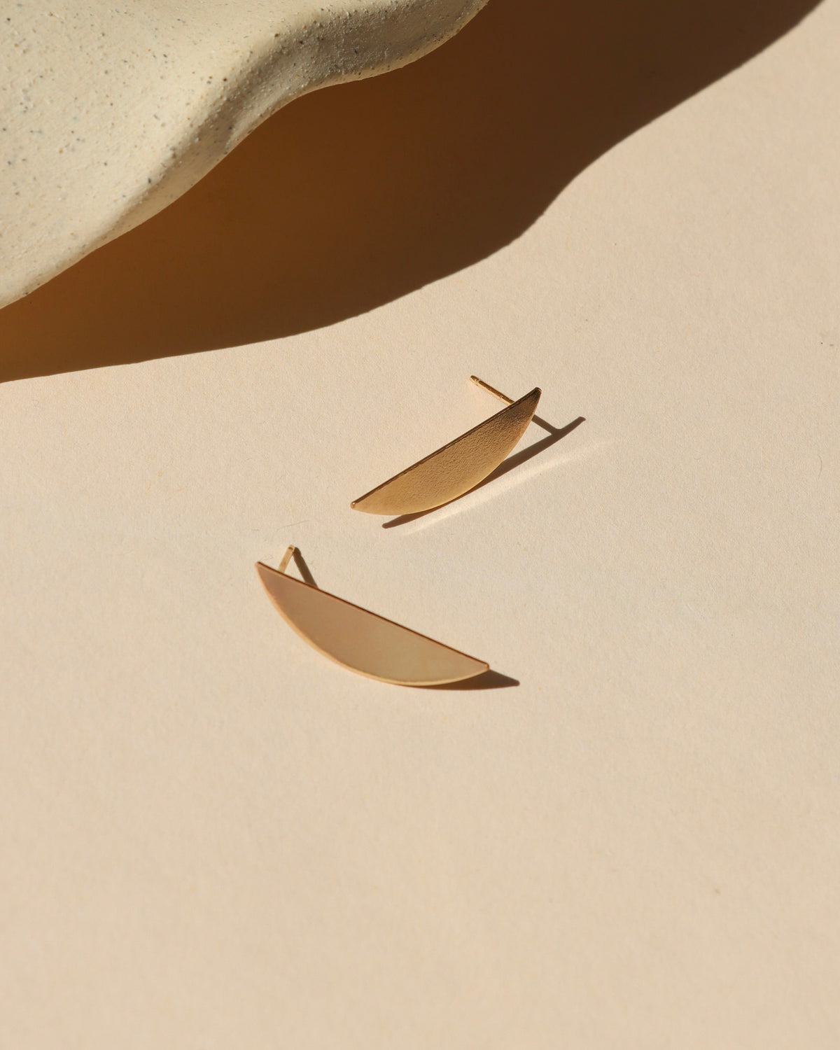 Pair of curved gold statement stud earrings photographed on a cream background in warm sunlight.