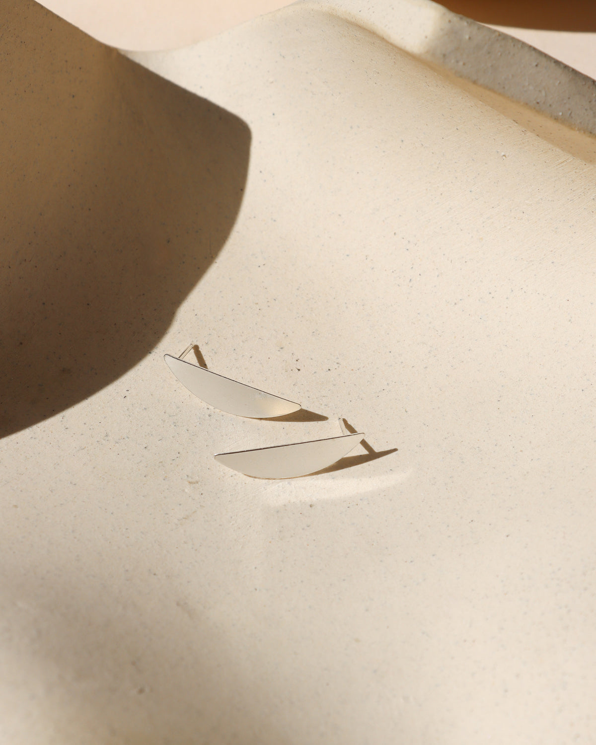 Pair of curved sterling silver statement stud earrings photographed on a cream background in warm sunlight.