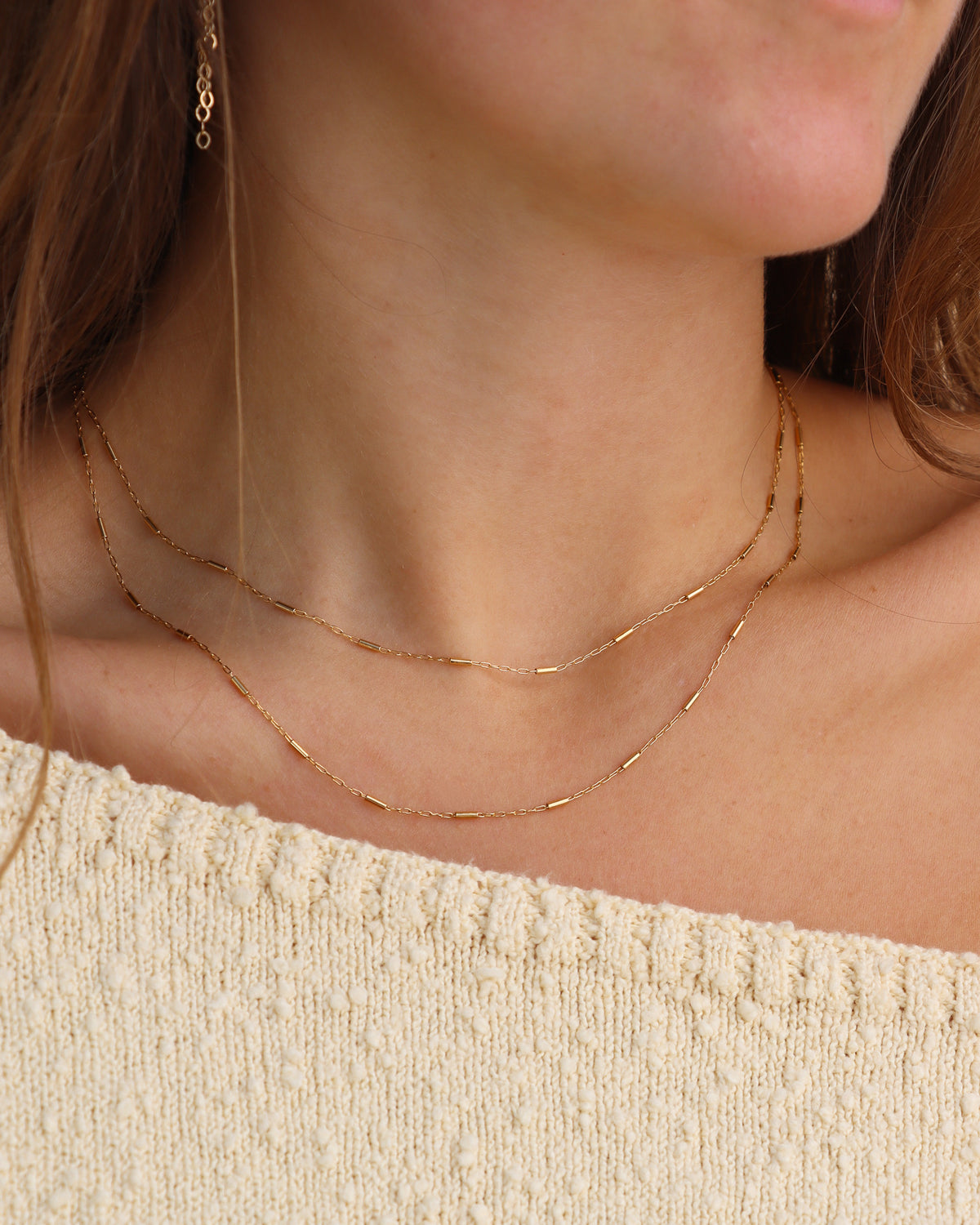 Close-up of a model wearing two layered gold Sola Necklaces at different lengths, featuring delicate chain links and small gold bar accents, styled with a cream knit sweater.