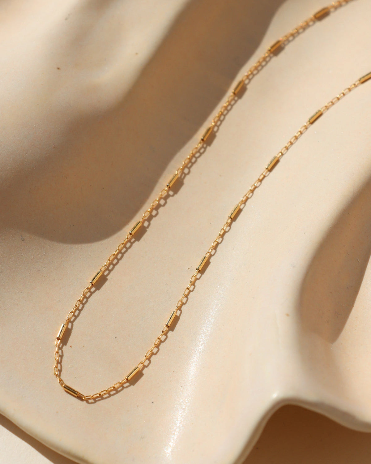 Delicate gold Sola Necklace featuring elongated chain links and evenly spaced polished gold bar accents, displayed on a cream ceramic surface in warm natural sunlight.