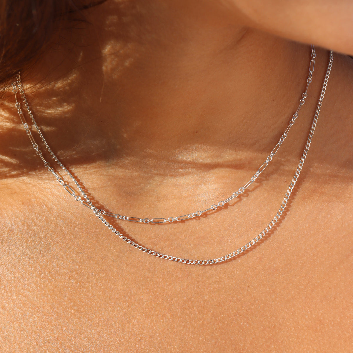 Extreme close-up shot of a woman's neckline highlighting two layered sterling silver chains in bright natural sunlight: a fine bar-and-link chain layered above a slightly longer delicate silver curb chain.