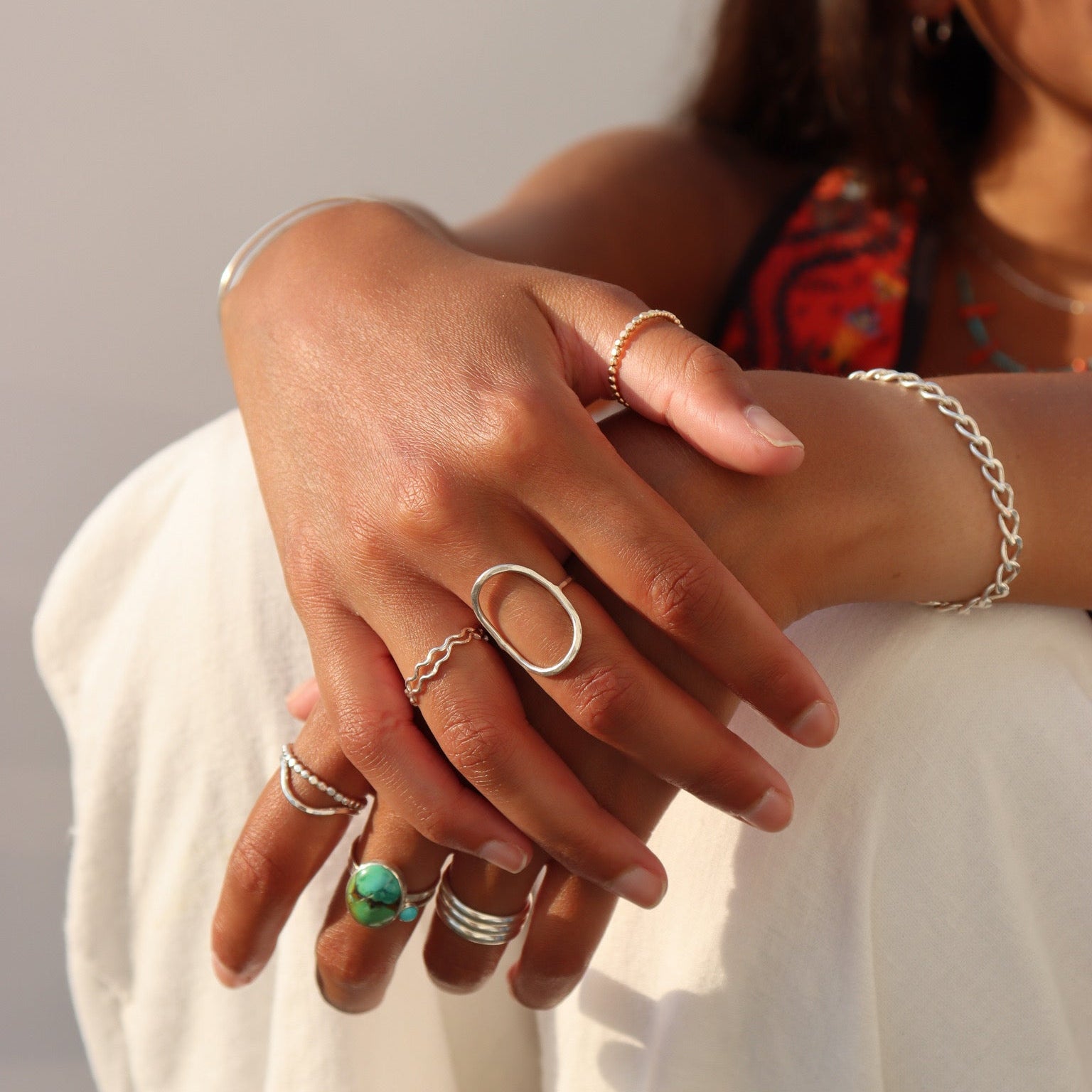 Lifestyle image of layered sterling silver rings, including a bold oval statement ring, natural turquoise ring, beaded stacking rings, and a wavy band paired with a sterling silver chain bracelet in warm natural sunlight.