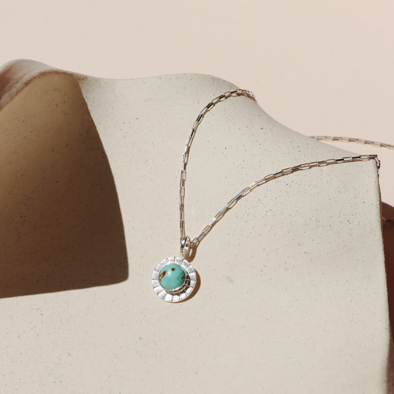 Silver paperclip-style chain necklace featuring a round turquoise stone with natural brown matrix, framed by a sunburst-textured bezel, resting on a speckled beige ceramic display under warm sunlight.