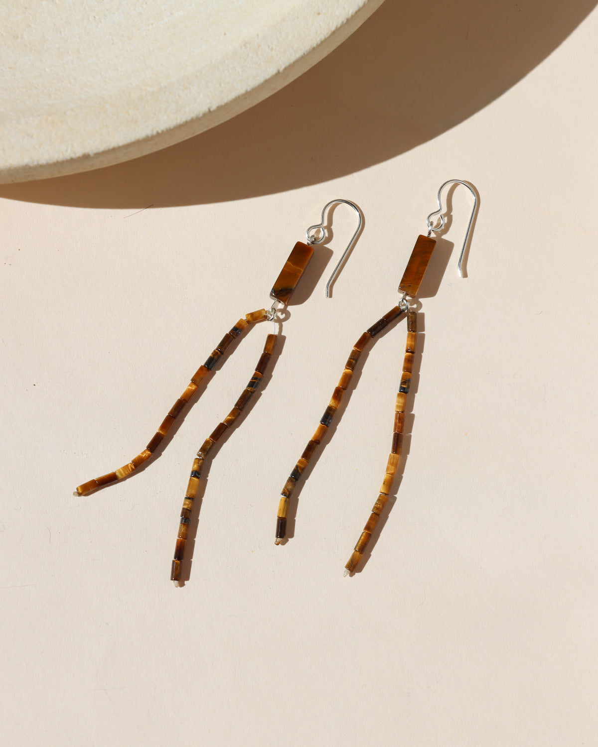 Long dangle earrings with rectangular tiger’s eye beads and strands of small brown and golden beads on silver ear wires, photographed in warm sunlight.