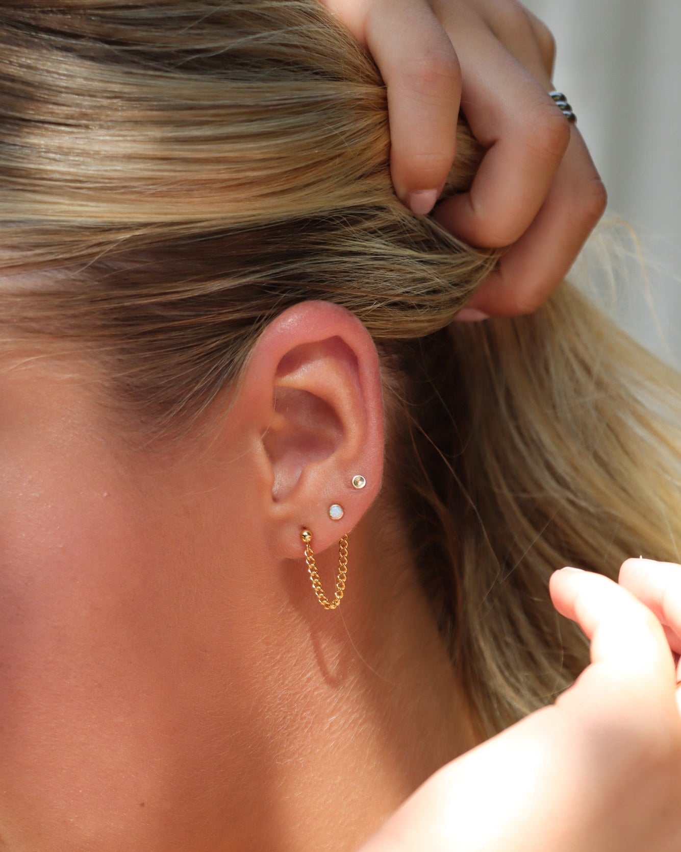 Close-up of a woman wearing layered gold earrings, including opal studs and a delicate chain earring, in warm natural light.