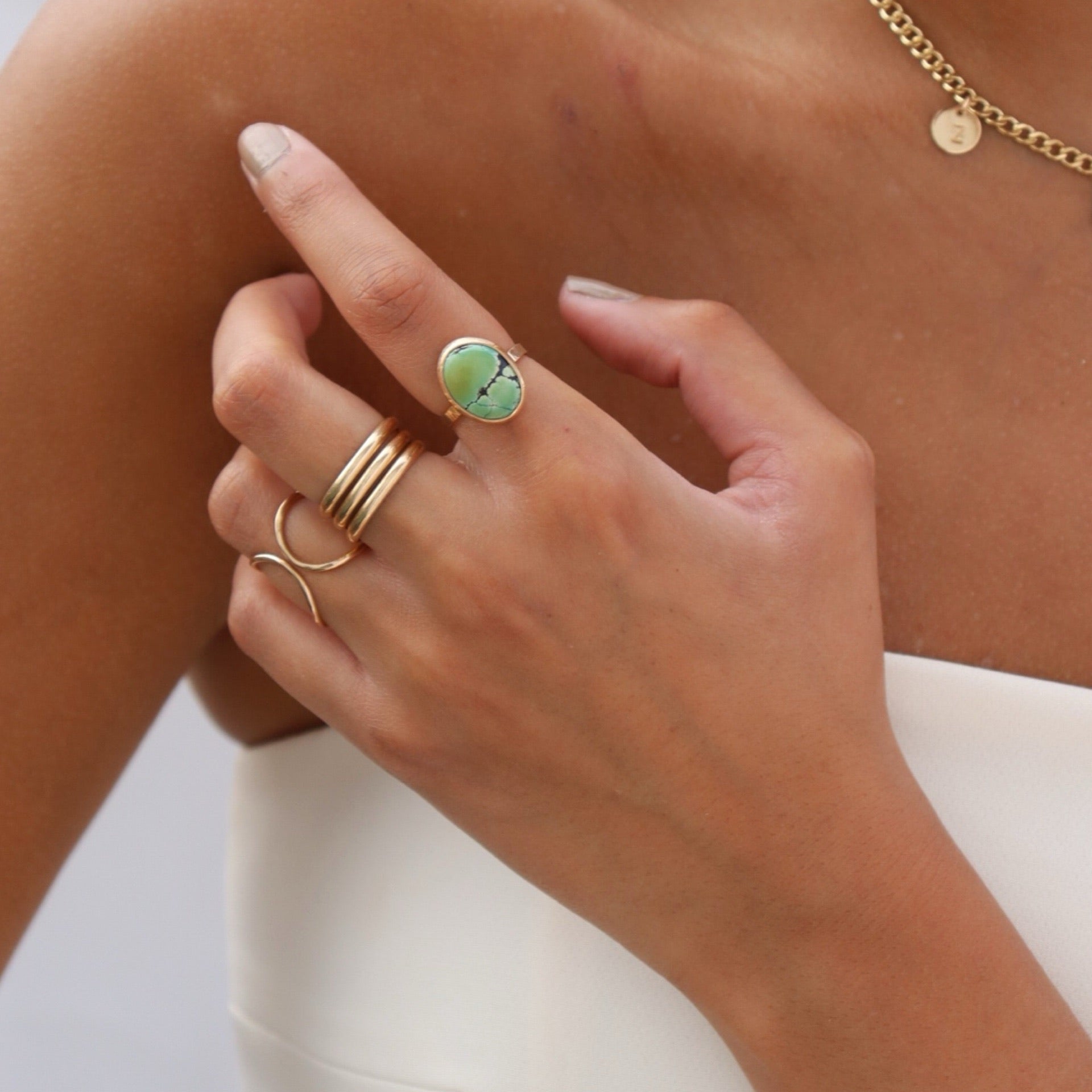 Close-up shot of a model's hand resting against her shoulder, displaying gold statement rings: an oval green turquoise gemstone ring on her index finger, a triple-stacked gold band on her middle finger, and an open double-arch ring on her ring finger.