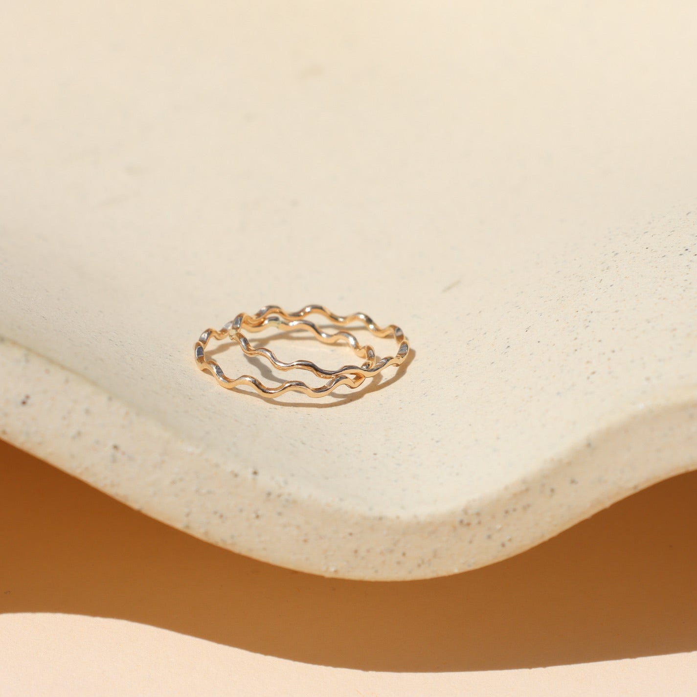 A pair of thin gold stackable rings with a squiggly wave design resting together inside a cream-colored speckled ceramic tray under warm sunlight.