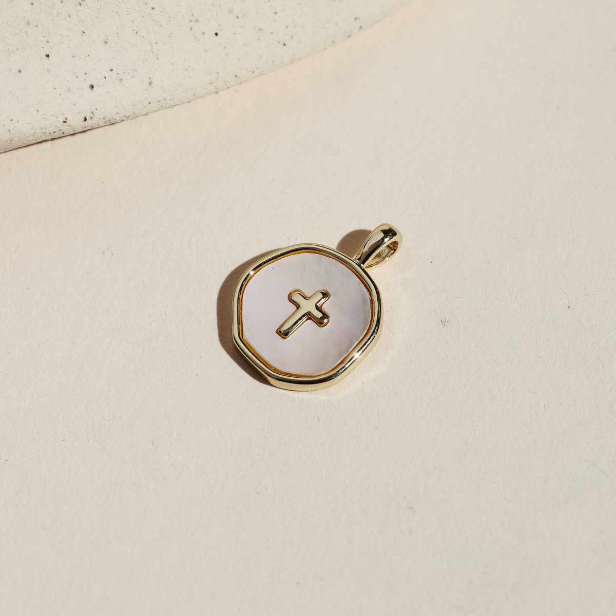 Round mother of pearl charm with a gold cross detail and gold-plated border, photographed on a neutral surface in warm natural light.