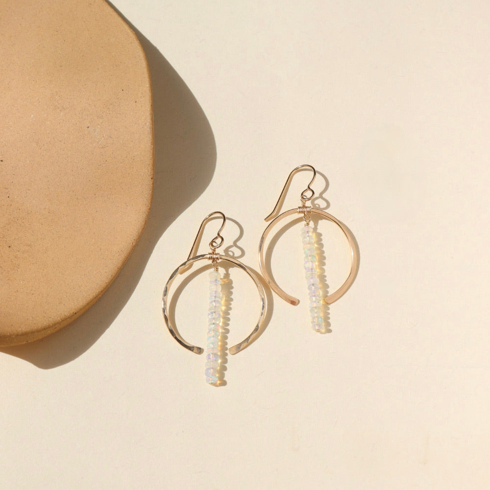 A 14k gold fill hoop featuring white opal gemstones dropped 2" from earring post. Earrings are displayed on a cream back drop in the sunlight. 