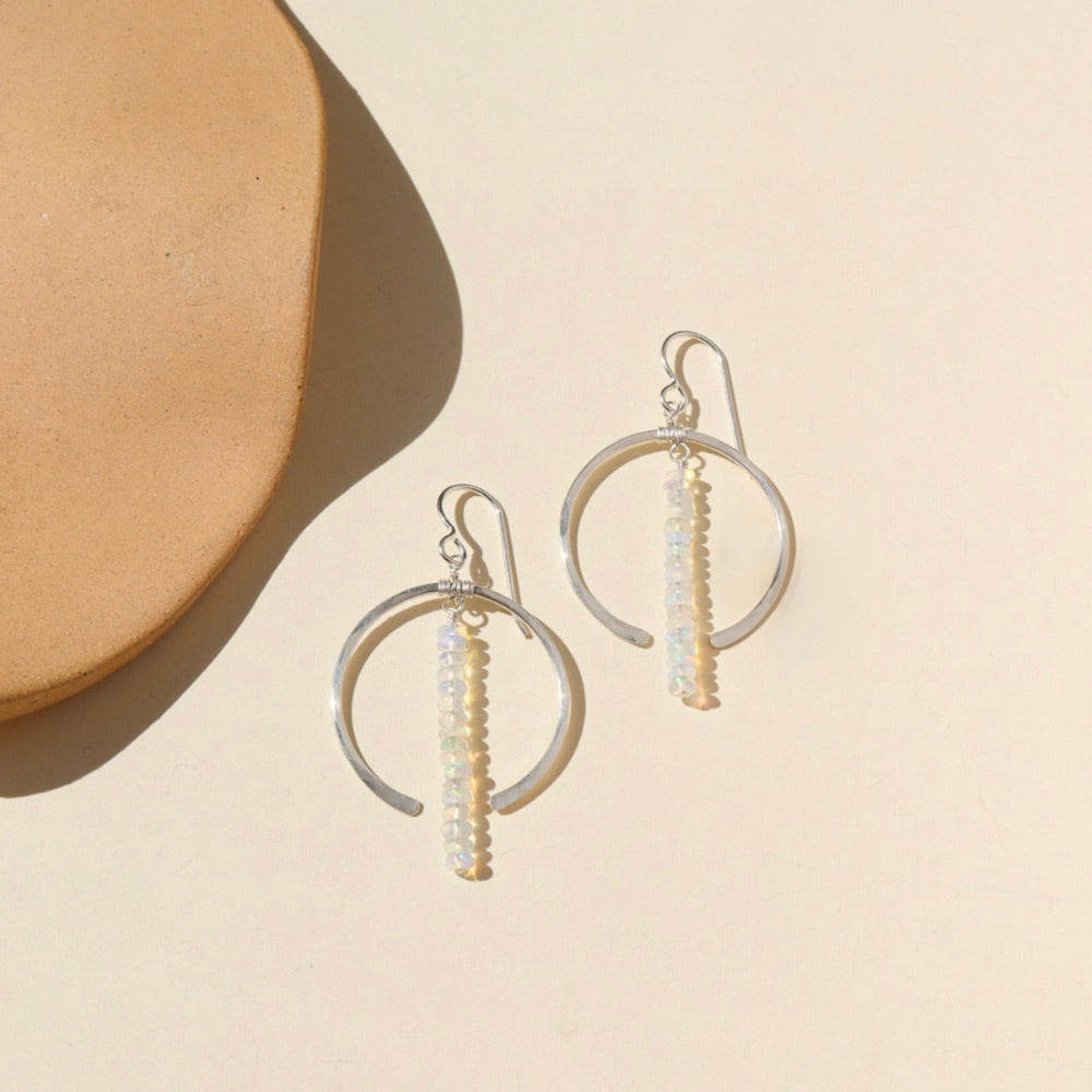 A sterling silver hoop featuring white opal gemstones dropped 2" from earring post. Earrings are displayed on a cream back drop in the sunlight. 
