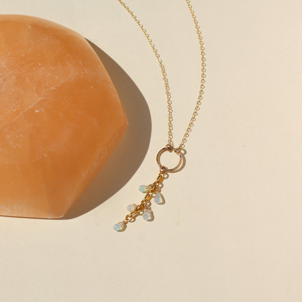 A 14k gold fill lariat with a standard chain and a 2" drop featuring 5 Teardrop Ethiopian Opals hanging from a rolo chain. The necklace is displayed on a cream back drop in the sunlight. 