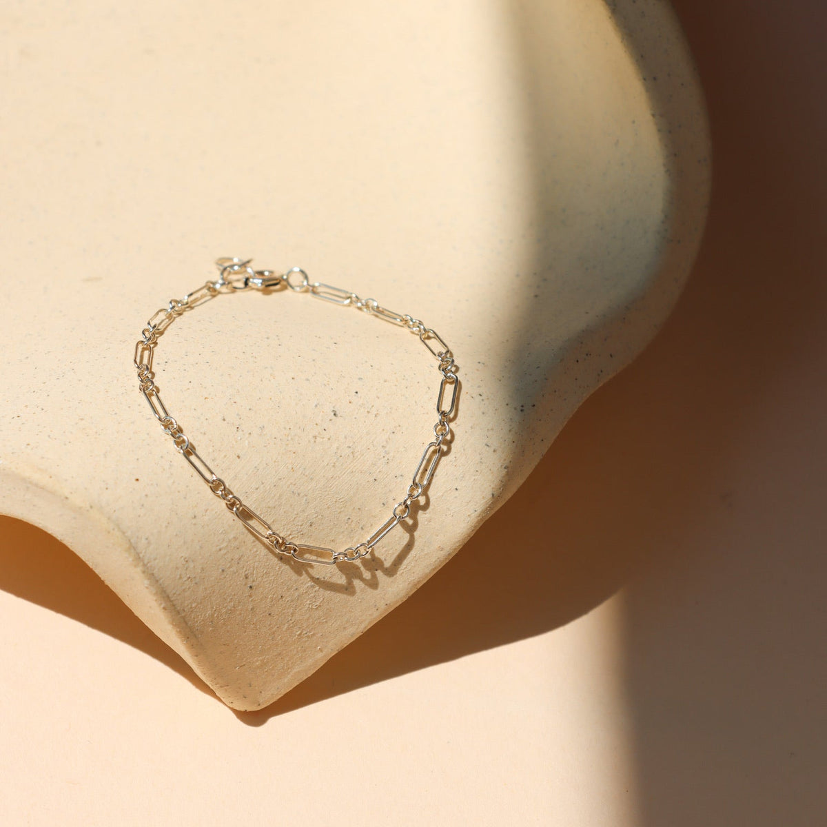 Minimalist fine silver paperclip link chain bracelet laid out on a beige speckled ceramic prop, bathed in soft, warm sunlight with subtle shadows.