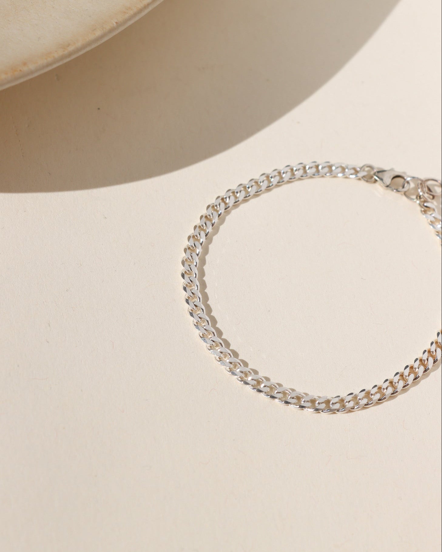 A silver curb chain bracelet with a lobster clasp, displayed on a warm beige surface with soft shadows.
