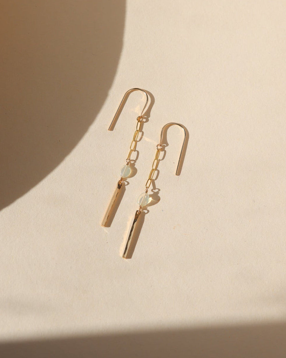 Gold drop earrings featuring delicate elongated chain links, luminous opal beads, and hammered gold bar pendants, photographed on a warm cream background with soft natural sunlight and shadows.