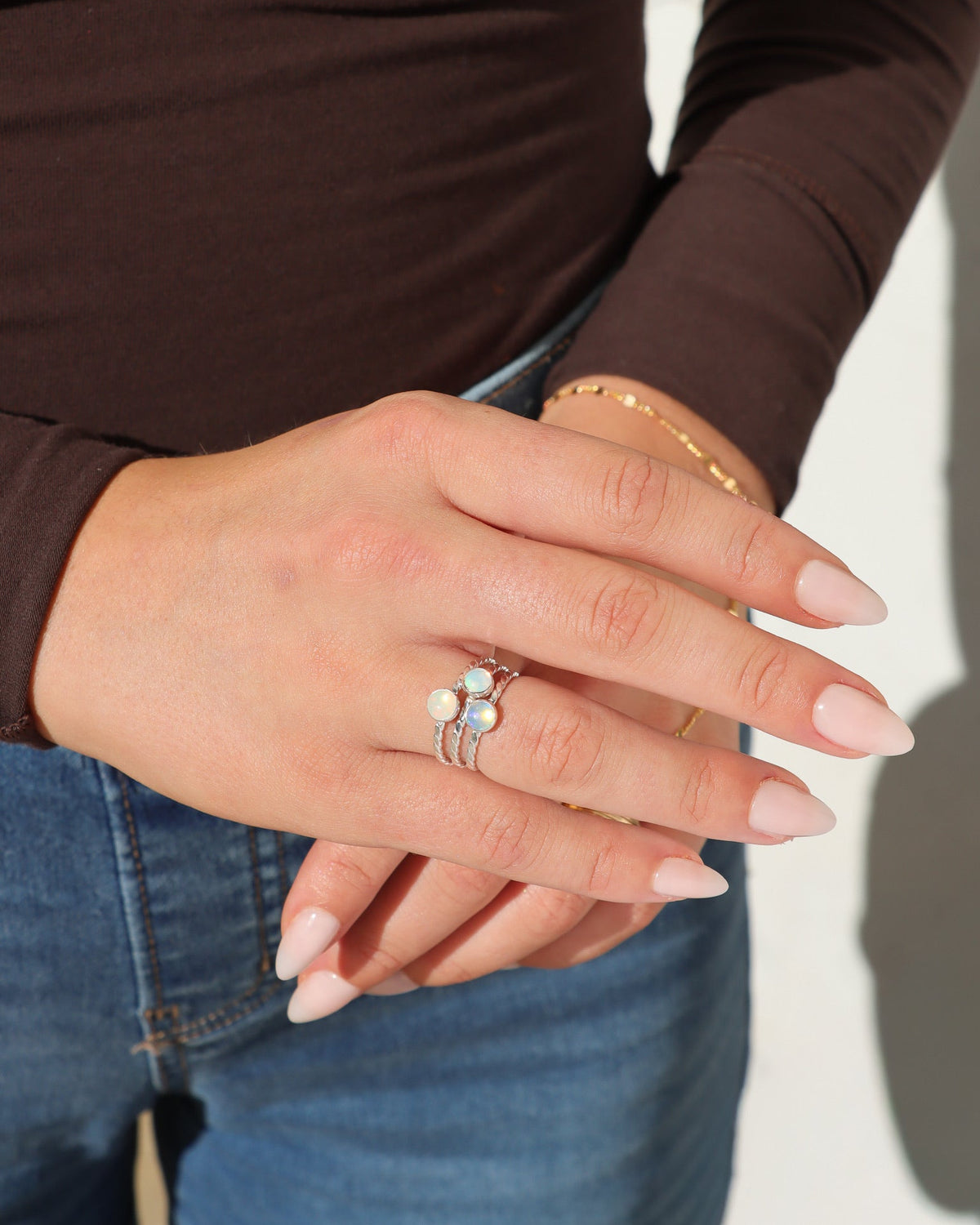 Model wearing three stacked silver twisted-band rings featuring luminous round opal gemstones, styled with a dark brown long-sleeve top, blue jeans, and a delicate gold bracelet in bright natural sunlight.