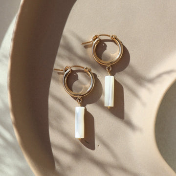 14k gold fill Mother of Pearl Hoops laid on a peach plate in the sunlight. These earring feature a classic hoop with a mother of pearl drop down stone. 