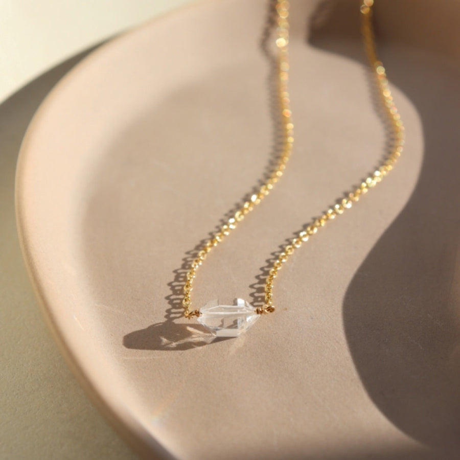 a clear double terminated quartz gemstone, known as a Herkimer Diamond, hangs from a delicate gold filled oval cable chain and is displayed on a ceramic jewelry dish