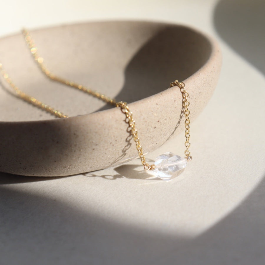 a clear double terminated quartz gemstone, known as a Herkimer Diamond, hangs from a delicate gold filled oval cable chain and is displayed on a ceramic jewelry dish